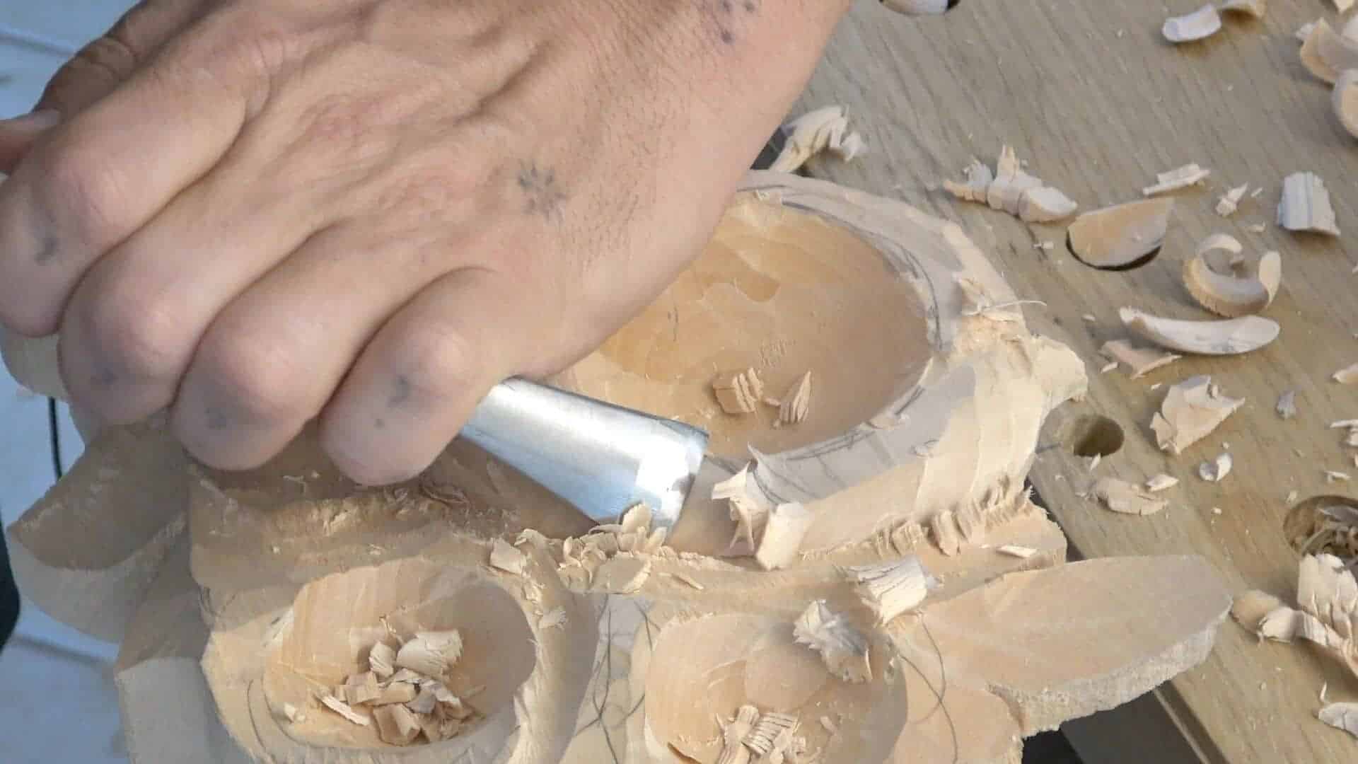 A person skillfully carving wood using a tool, focusing on creating details. Shavings scatter around on the workbench made of light-colored wood. This scene perfectly captures the hands-on experience of wood carving classes and lessons.