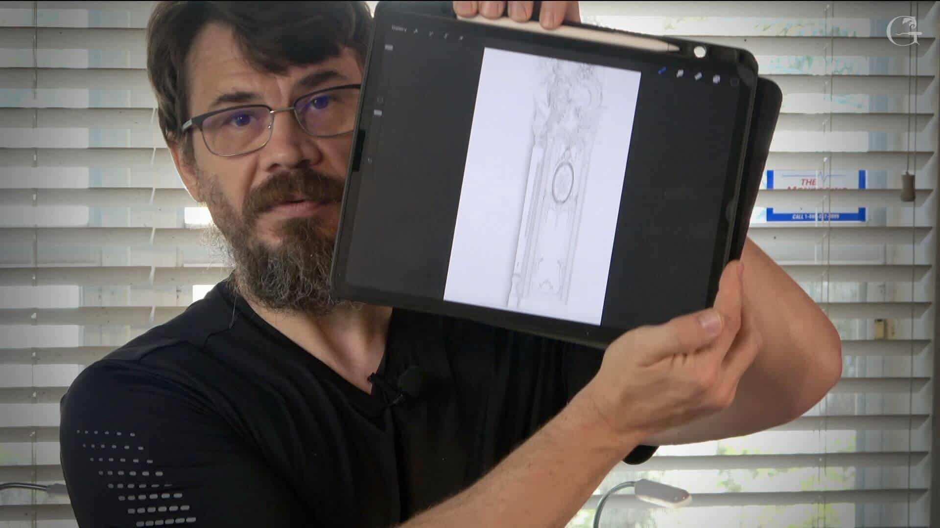 A person showing a drawing of a wood carving design on a tablet. The setting features blinds and an interesting workspace. Perfect for anyone interested in wood carving classes or lessons!