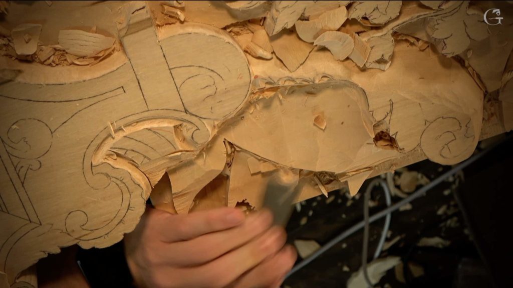 A close-up of a hand working on a wood carving project. The person is using a chisel to shape the wood, which shows detailed patterns outlined on the surface. Shavings and wood chips are scattered around, hinting at the carving process. This image perfectly captures the dedication and skill involved in wood carving classes and lessons.