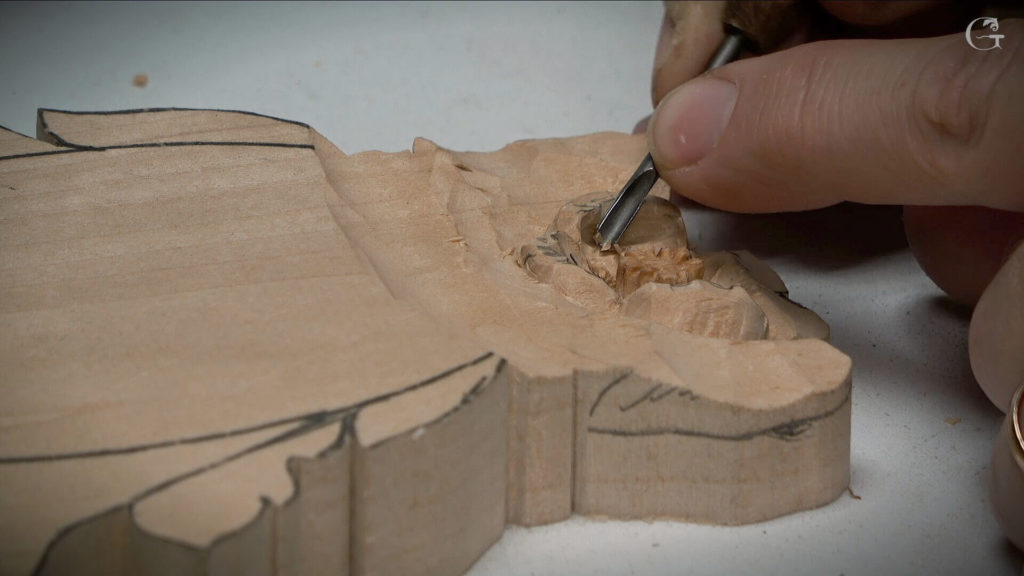A close-up of a hand carving a wooden piece using a chisel. The wood appears to be light-colored, likely basswood, with pencil lines marking the design on top. This image captures the careful and detailed work involved in wood carving, showcasing a specific moment in a wood carving class or lesson.
