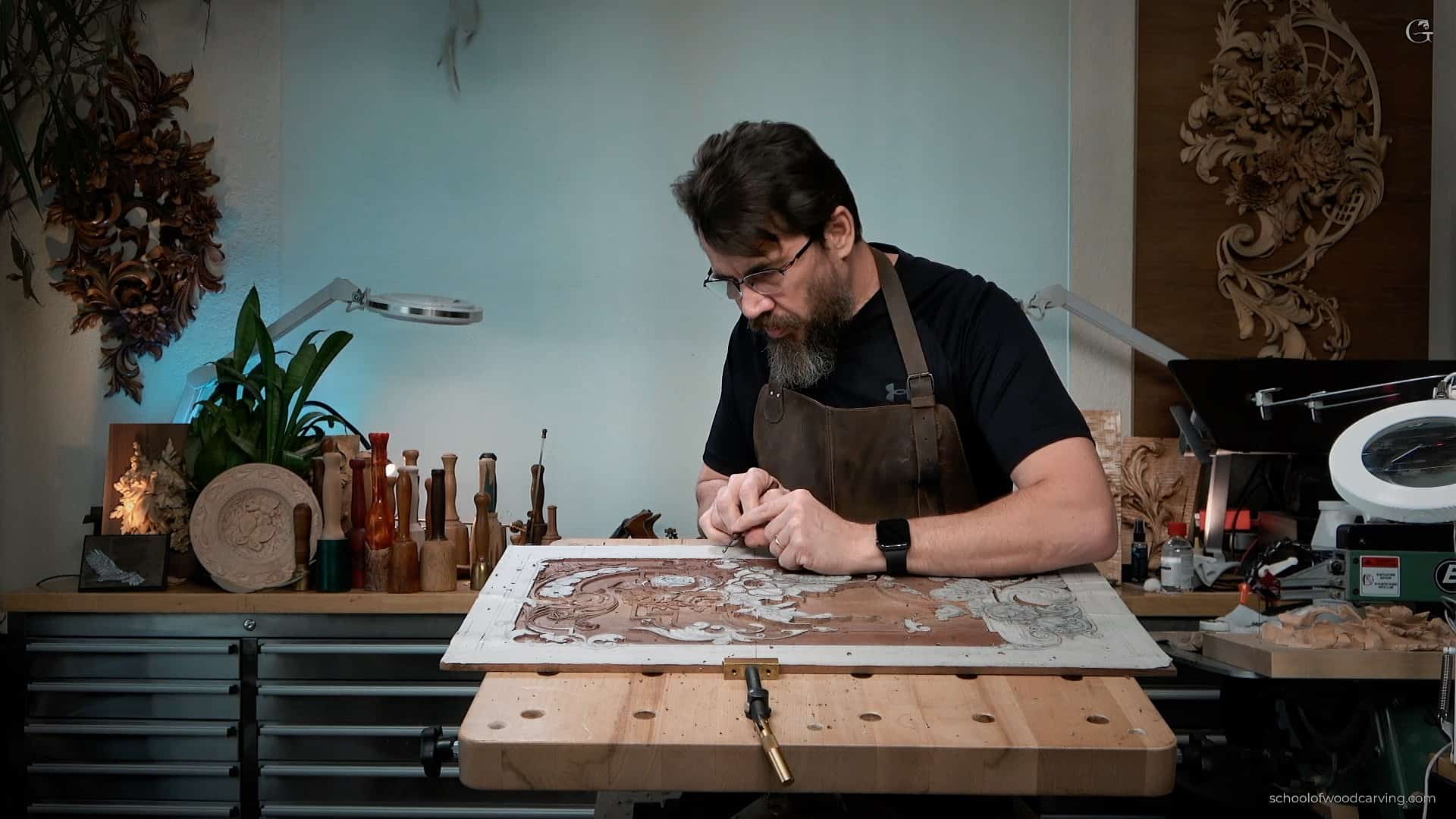 A wood carver intensely focused on his work at a carving bench. Surrounding him are various tools and carving pieces, including intricate designs and finished projects. The workshop has a warm, creative atmosphere, perfect for wood carving classes and lessons.