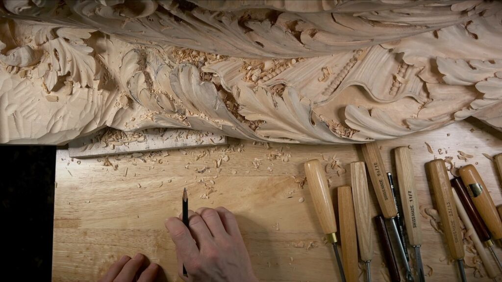 A pair of hands working on intricate wood carving, with woodworking tools and wood shavings spread out on a wooden surface, reminiscent of the techniques found in woodcarving tutorials online.